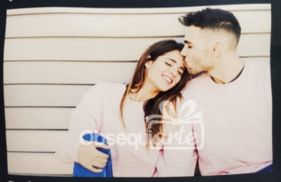 Un hombre besa la frente de una mujer mientras están sentados juntos, ambos visten blusas de colores claros y sobre un fondo de rayas.